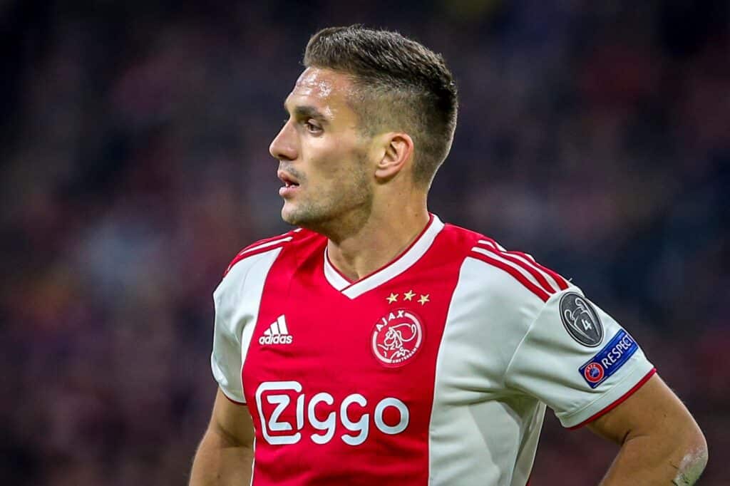 Plano aproximado de perfil de um jogador de futebol com a camisola principal do Ajax de Amesterdão, que apresenta as cores distintivas vermelho e branco e o logótipo do patrocinador Ziggo. O jogador usa um patch da Liga dos Campeões UEFA na manga e aparenta estar a transpirar durante um jogo. A camisola também exibe as icónicas três estrelas acima do emblema do Ajax, representando os múltiplos campeonatos do clube. O jogador tem um corte de cabelo curto e moderno e está a olhar para o lado